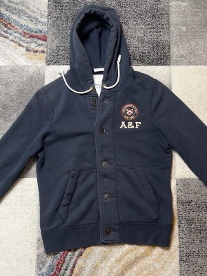 Mörkblå hoodie från Abercrombie & Fitch - Mörkblå hoodie med knappar framtill och vit dragsko i huvan. Broderad A&F-logga och emblem på bröstet. Två fickor fram och ribbade muddar. Tillverkad i mjuk bomullsblandning för en skön känsla.