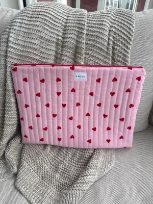 Rosa datorväska med röda hjärtan - Supersöt datorväska från EMELO i quiltad rosa textil med små röda hjärtan över hela väskan. Väskan har dragkedja upptill och en mjuk, vadderad känsla som skyddar din laptop. Perfekt för dig som vill ha en gullig och unik look till din dator. Passar maxmått på laptop: 33x24 cm