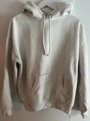 Vit hoodie med ficka - En enkel och stilren vit hoodie med stor känguruficka framtill och justerbar dragsko i huvan. Hoodien har ribbade muddar vid ärmslut och nederkant. Perfekt för en avslappnad och bekväm look. Materialet känns mjukt och skönt mot huden.