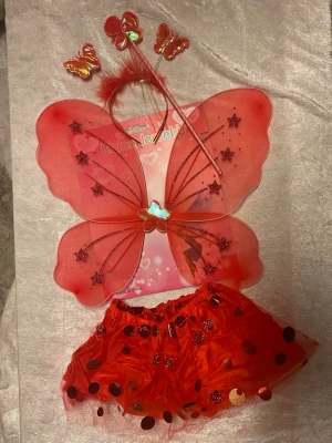 Barn Rött fjäril maskeradset med kjol - Säljer ett rött maskeradset med fjärilstema som innehåller vingar med glitterstjärnor, matchande kjol med stora paljetter, diadem med fjärilar och en trollstav. Perfekt för maskerad eller cosplay. Materialet är lätt mesh och plast.