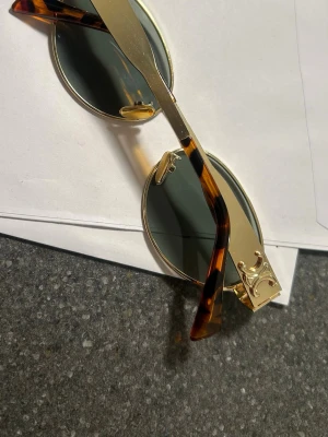 Celine solglasögon guld - Säljer ett par runda solglasögon från Celine med guldfärgade metallbågar och sköldpaddsmönstrade skalmar. Glasen är mörka och bågarna har snygga gulddetaljer vid gångjärnen. Perfekt accessoar för dig som vill ha en lyxig och trendig look.