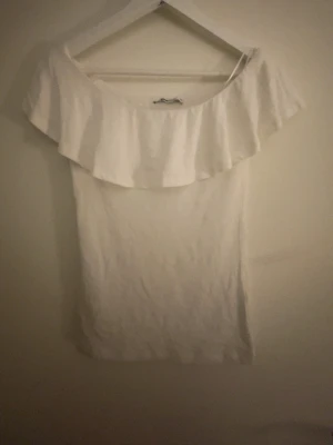 Vit offshoulder topp med volang - Säljer en vit offshoulder topp med bred volang runt axlarna. Toppen är tight och har en enkel, stilren design som passar perfekt till jeans eller kjol. Materialet känns mjukt och stretchigt, vilket gör den bekväm att bära.