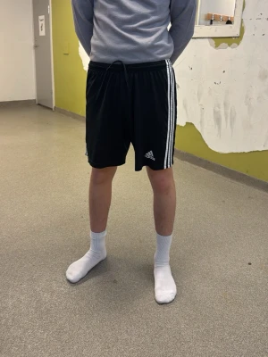 Svarta Adidas shorts med vita ränder - Säljer ett par klassiska svarta Adidas shorts med vita ränder längs sidorna och logga framtill. Shortsen har snörning i midjan och är perfekta för träning eller chill. Bekväma och sportiga med normal passform. En bra dasse behövs 