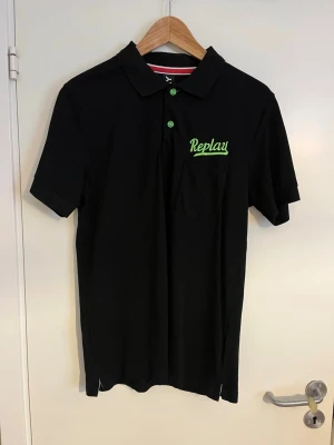 Svart pikétröja från Replay - Snygg svart pikétröja från Replay med grön logga broderad på bröstet och matchande gröna knappar. Klassisk krage och korta ärmar, perfekt för en avslappnad men stilren look.
