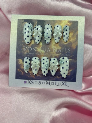 DollyDots (XS);Medium Almond - Handgjorda press-on naglar i vit bas med mörkblåa prickar, dekorerade med små 3D-rosetter och söta blåa pärlor. Naglarna har en medium almondform och är perfekta för dig som vill ha en unik och lekfull look. XS: 14 mm, 11 mm, 11,5 mm, 10 mm, 8 mm 