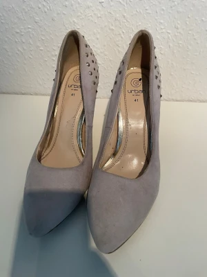 Ljusgrå pumps med nitar från Urban - Snygga ljusgrå pumps från Urban med spetsig tå och klack. Skorna är dekorerade med silverfärgade nitar på hälen och har en ovandel i mockaimitation. Perfekta för dig som vill ha en edgy men stilren look.