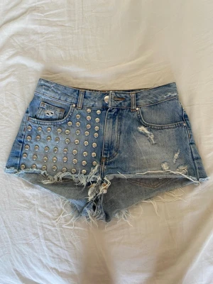 Blå jeansshorts med nitar från Bik Bok - Säljer ett par blå jeansshorts från Bik Bok med råa kanter och slitningar. Ena sidan är dekorerad med silvriga nitar för en edgy look. Klassisk femficksmodel. Perfekta för sommaren!