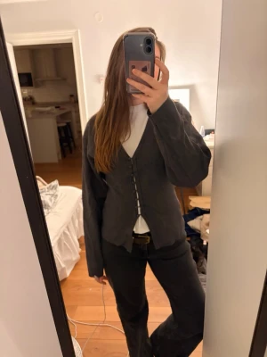 Mörkgrå kofta - Säljer en mörkgrå kofta med v-ringning och knappar framtill. Koftan har långa ärmar och en avslappnad passform som ger en cool och trendig look. Perfekt att bära över en t-shirt eller polotröja för en lager-på-lager stil.