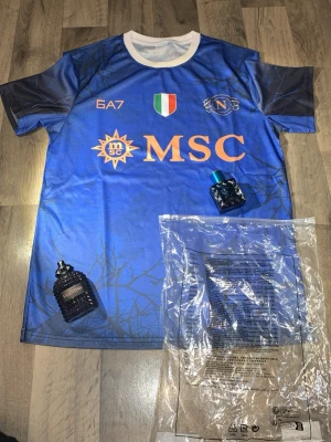 Blå Napoli matchtröja GA7 - Snygg blå matchtröja från Napoli med vita och orange detaljer. Framsidan har MSC-logga, klubbmärke och italienska flaggan, medan baksidan har SORGESANA-tryck. Tillverkad i lätt och ventilerande syntetmaterial, perfekt för fotboll eller träning.