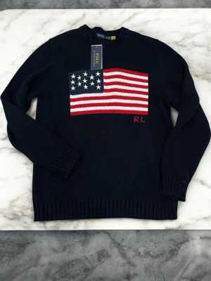 Svart stickad tröja Polo Ralph Lauren - Stickad svart tröja från Polo Ralph Lauren med amerikansk flagga i rött, vitt och blått på bröstet samt RL-broderi. Tröjan har rund hals, långa ärmar och ribbade muddar. Perfekt för dig som gillar klassisk och stilren design.