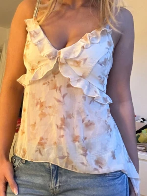 Blommig volangtop med knyt i ryggen - Söt blommig linnetopp i ljusa toner med volangdetaljer framtill och tunna axelband. Toppen har öppen rygg med knyt och är tillverkad i ett tunt, luftigt material som känns som polyester eller liknande. Perfekt för varma dagar och en avslappnad look.