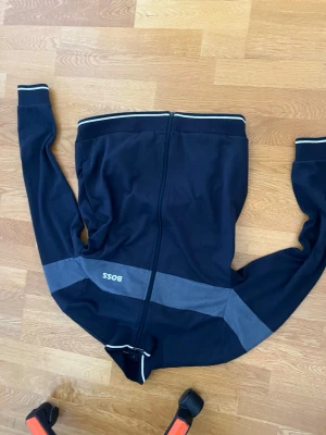 Marinblå zip-hoodie från BOSS - Snygg marinblå hoodie från BOSS med dragkedja framtill och en bred grå rand över bröstet och armarna. Tröjan har vita detaljer på muddar och krage samt BOSS-logga på bröstet. Perfekt för en avslappnad och sportig stil.