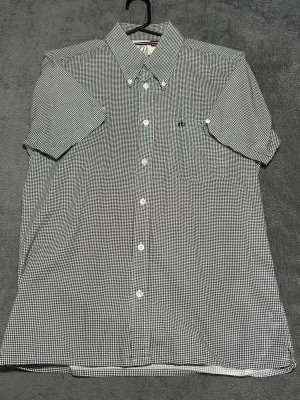 Merc London Shirt – Houndstooth – Short Sleeve – Size S - Kortärmad skjorta från Merc London i klassiskt houndstooth-mönster.  Perfekt för en stilren mod / british look. Lätt att matcha och funkar både casual och uppklätt.  Detaljer • Merc London • Kortärmad • Houndstooth (svart/vit) • Button down • Storlek S • 100% bomull  📦 Skickas snabbt 💬 Skriv vid frågor
