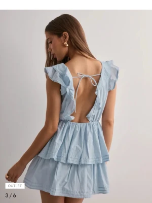 Ljusblå volangklänning med öppen rygg - Söt ljusblå kortklänning med volangdetaljer och omlott framtill. Klänningen har ärmlös design, öppen rygg med knytband och dubbla lager i kjolen för extra volym. Perfekt för sommarens alla tillfällen.