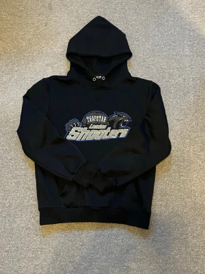 Svart Trapstar London hoodie - Snygg svart hoodie från Trapstar London med stort tryck på bröstet där det står 'Shooters' i silver och blått. Klassisk huva med dragsko och ribbade muddar vid ärmar och midja. Perfekt för en streetwear-look.