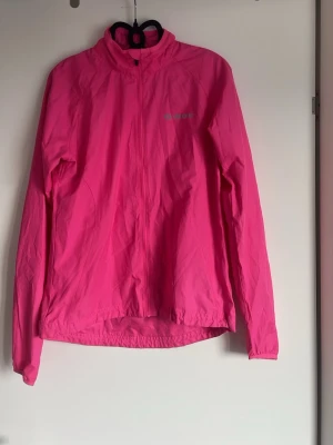 Rosa vindjacka från soc - Snygg rosa vindjacka från soc med hög krage och hel dragkedja framtill. Jackan har långa ärmar, reflekterande detaljer och lätt, vindtätt material som passar perfekt för löpning eller cykling. Enkel och sportig design med logga på bröstet. Den är knappt använd 