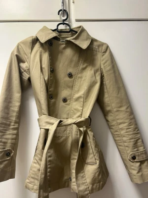 Beige trenchcoat från G-Star Raw - Stilren beige trenchcoat från G-Star Raw med dubbelknäppning, knytskärp i midjan och klassisk krage. Jackan har två fickor framtill och dekorativa knappar vid ärmsluten. Materialet är kraftigt och ger en snygg siluett.