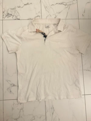 Burberry Pique - Klassisk vit pikétröja från Burberry med korta ärmar och krage. Tröjan har diskret broderad logga på bröstet och ikoniskt rutigt tyg på insidan av knappslån. Perfekt till en clean och stilren look.