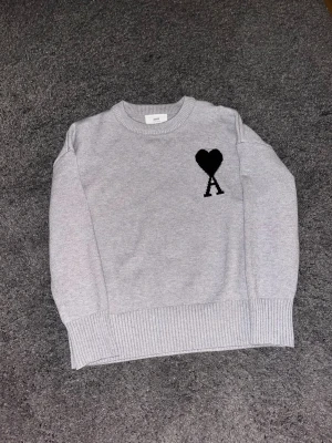 Grå stickad tröja från Ami - Grå stickad tröja från Ami med svart hjärta och bokstaven A broderat på bröstet. Tröjan har rund halsringning, ribbade muddar och är tillverkad i mjukt material. Perfekt för en stilren och trendig look.
