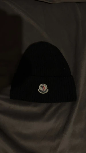 Svart Moncler mössa i ribbstickad ull - Svart ribbstickad mössa från Moncler med klassisk uppvikt kant och broderad logotyp framtill. Tillverkad i mjuk ull som håller dig varm och stilren under kalla dagar. Perfekt accessoar för att lyfta din vinterlook.