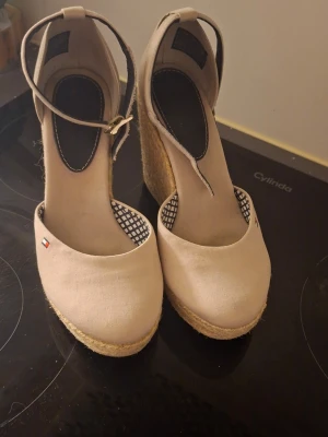 Beige kilklackade sandaler Tommy Hilfiger - Snygga beige sandaler från Tommy Hilfiger med hög kilklack i flätad jute och rund tå. Skorna har justerbart ankelband med spänne och diskret logga på sidan. Ovandelen är i textil och innersulan har ett svartvitt rutmönster.