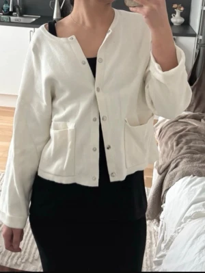 Vit kofta med knappar och fickor - Säljer en stilren vit kofta med knäppning framtill och två stora fickor. Koftan har en rak passform och långa ärmar. Perfekt att slänga över en topp för en enkel och clean look.