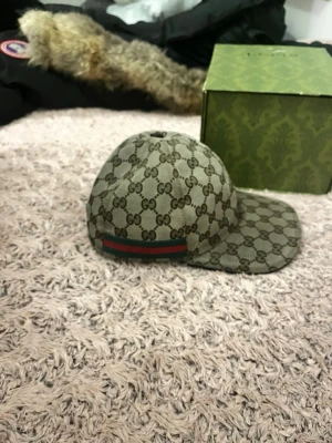 Gucci beige GG Supreme keps - Säljer en äkta  beige keps från Gucci med klassiskt GG Supreme-mönster över hela kepsen och en grön-röd rand baktill. Kepsen har böjd skärm och är tillverkad i canvasmaterial. Box och dustbag från Gucci medföljer .