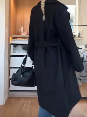 Svart lång kappa med skärp - Stilren svart lång kappa med skärp i midjan och bred krage. Klassisk rak passform som ger en snygg siluett. Perfekt för kyligare dagar och enkel att matcha med olika outfits.