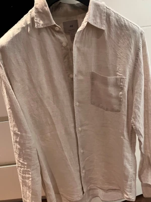 Vit linneskjorta från H&M - Säljer en stilren vit linne skjorta från H&M i regular fit. Skjortan är tillverkad i linne, har lång ärm, klassisk krage, bröstficka och knäppning framtill. Perfekt för en avslappnad men ändå snygg look.