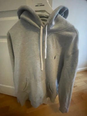 Grå hoodie från Polo Ralph Lauren - Snygg grå hoodie från Polo Ralph Lauren med klassisk huva och vita dragsnören. Har en känguruficka framtill och den ikoniska lilla blå loggan broderad på bröstet. Tillverkad i mjukt bomullsmaterial som är perfekt för chill dagar.