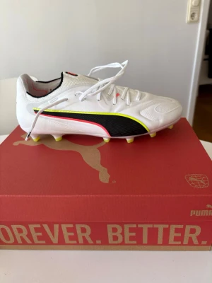 Vita Puma fotbollsskor elite fg - Puma kings, Snygga vita fotbollsskor från Puma med klassisk svart logga på sidan, gula och röda detaljer samt vita skosnören. Skorna har fasta dobbar i gult och är designade för spel på gräsplan. Ovandelen är i syntetmaterial för bra passform och lätt vikt. Storlek 44.5 EU , endast testade, påse ingår