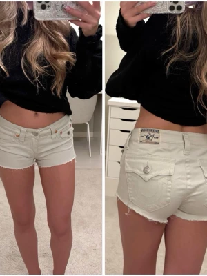 Vita denimshorts från True Religion - Säljer ett par vita denimshorts från True Religion med råa kanter och klassiska fickor bak. Shortsen har låg midja, knappgylf och detaljerade sömmar. Perfekta för sommaren och riktigt snygga till en croppad topp.