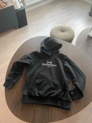 Svart hoodie Peak Performance - Svart hoodie från Peak Performance med vit logga och text på bröstet. Mjuk och skön, perfekt för en avslappnad stil. Huva utan dragsko.