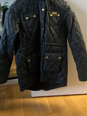 Svart jacka från Barbour - Snygg svart jacka från Barbour med gulddetaljer och fyra fickor framtill. Jackan har huva med fuskpäls och tryckknappar. 