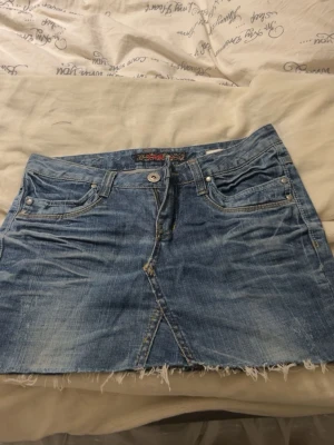 Blå jeanskjol - Säljer en kort blå jeanskjol med fransig, rå kant nertill och klassiska fem fickor. Kjolen har knapp och dragkedja framtill samt slitna detaljer för en avslappnad look. Perfekt till sommaren och enkel att matcha med olika toppar. Ingen storlek på de men passar xs-s❤️