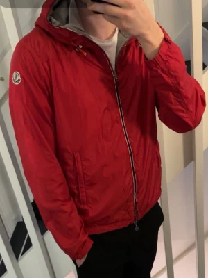 Moncler urville jacka  - Snygg röd vindjacka från Moncler med huva och grått innerfoder. Jackan har dragkedja framtill, två sidofickor och Moncler-logga på ärmen. Perfekt för blåsiga dagar och enkel att matcha till olika outfits. 