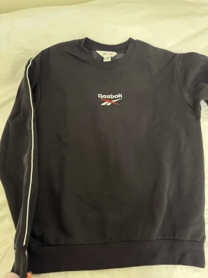Svart sweatshirt från Reebok - Svart sweatshirt från Reebok med vit piping längs ärmarna och broderad logga på bröstet i vitt och rött. Klassisk rund halsringning och ribbade muddar vid ärmslut och nederkant. Perfekt för en sportig och avslappnad stil.