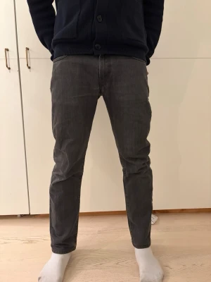 J.Lindeberg Jeans - Säljer ett par gråa jeans med rak passform och klassiska fickor både fram och bak. Byxorna har diskreta ränder i tyget och är perfekta för dig som gillar en stilren och avslappnad look. Materialet är mjukt och bekvämt, perfekt för vardagsoutfits.