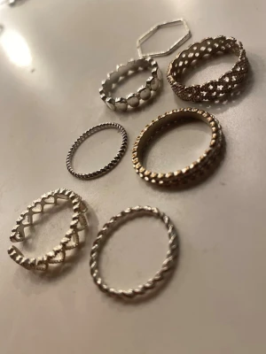Sju stilrena ringar i silver och guld - Säljer ett set med sju ringar i olika former och mönster, både i silver- och guldfärg. Ringarna har detaljer som hjärtan, pärlstruktur, tvinnad design och en sexkantig form. Perfekta för att mixa och matcha för en personlig stil. 10kr/st, alla 7st för 50kr!