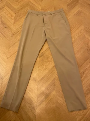 Beige kostymbyxa med raka ben - Snygga beige kostymbyxor med raka ben och pressveck. Byxorna har bälteshällor, sidofickor fram och två passpoalfickor bak. Perfekta för en clean och stilren look. Materialet ser ut att vara en mjuk och lätt blandning, troligtvis polyester eller bomull.