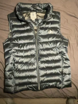 Grå dunväst från U.S. Polo Assn - Snygg grå dunväst från U.S. Polo Assn med quiltad design och hög krage. Västen har dragkedja framtill och en logga på bröstet. Perfekt för lager på lager och ger en sportig vibe till din outfit.