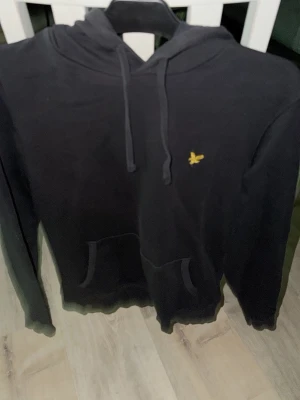 Svart hoodie från Lyle & Scott - Svart hoodie från Lyle & Scott med klassisk gul logga på bröstet. Hoodien har huva med dragsko, magficka och ribbade muddar. Tillverkad i mjukt bomullsmaterial som är skönt att bära. Perfekt för en avslappnad och stilren look.