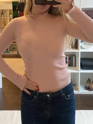 Ljusrosa ribbad stickad polotröja - Supersöt ljusrosa stickad tröja med ribbad struktur och lång ärm. Tröjan har en hög hals med volangkant som ger en extra detalj. Figurnära passform och mjukt material med vida ärmar.💕