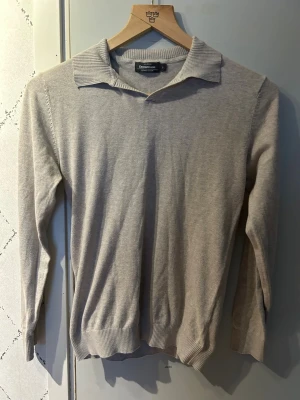 Beige stickad tröja med krage - Stilren beige stickad tröja från Dressmann med klassisk krage och långa ärmar. Tröjan har ribbade muddar vid ärmslut och nederkant, vilket ger en snygg passform. Perfekt för dig som gillar en enkel men elegant look.