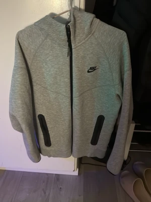 Grå Nike tech helt ny - Snygg grå hoodie från Nike med svart logga på bröstet och hel dragkedja framtill. Två svarta fickor med dragkedja och huva för extra komfort. Perfekt för chill dagar eller när du vill ha en sportig look.