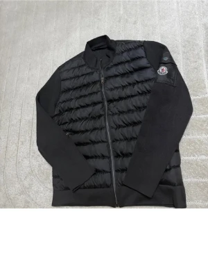 Svart cardigan från Moncler - Snygg svart pufferjacka från Moncler med quiltad front och släta ärmar. Jackan har dragkedja framtill, ribbad krage och Moncler-logga på ärmen. Materialet är en mix av blankt nylon och mjukare tyg på ärmarna. Perfekt för kalla dagar.