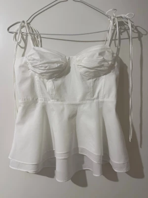 Zara Vit korsettopp med knytband - Supersöt vit korsettopp med tunna axelband som knyts på axlarna. Toppen har markerade kupor och en lätt utsvängd nederdel som ger en romantisk vibe. Perfekt att styla med jeans eller kjol för en trendig look.