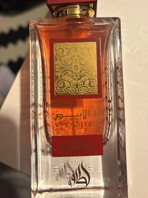 Lattafa Ana Abiyedh Scarlet parfym - Ana Abiyedh Scarlet från Lattafa är en parfym i en elegant, rektangulär glasflaska med röd-orange doftvätska. Flaskan har gulddetaljer och orientaliskt mönster på etiketten. Texten är i både arabiska och engelska, vilket ger en lyxig och unik känsla.
