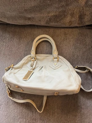 Ljusgrå Marc by Marc Jacobs handväska - Snygg ljusgrå handväska från Marc by Marc Jacobs i mjukt läder. Väskan har både handtag och axelrem, guldfärgade metalldetaljer och en dekorativ tag. Perfekt för dig som gillar stilrena och klassiska accessoarer.