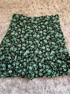 Grön blommig kortkjol med volang - Supersöt kortkjol med grönt blommönster och volangkant. Kjolen har en bekväm passform och är perfekt för dig som vill ha en lekfull och trendig stil. Passar till både sneakers och boots för en cool look.